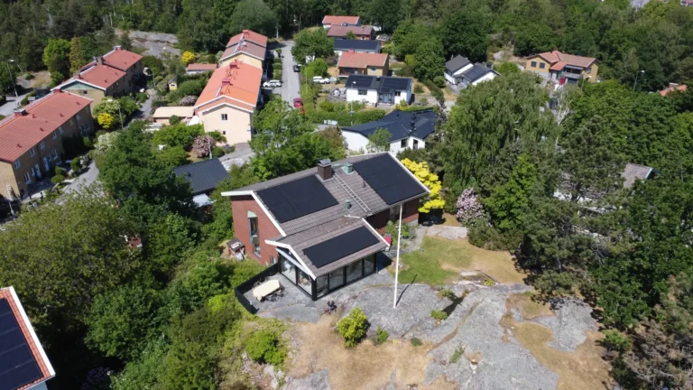Svarta solpaneler på sadeltaket av en röd tegelvilla omgiven av natur.