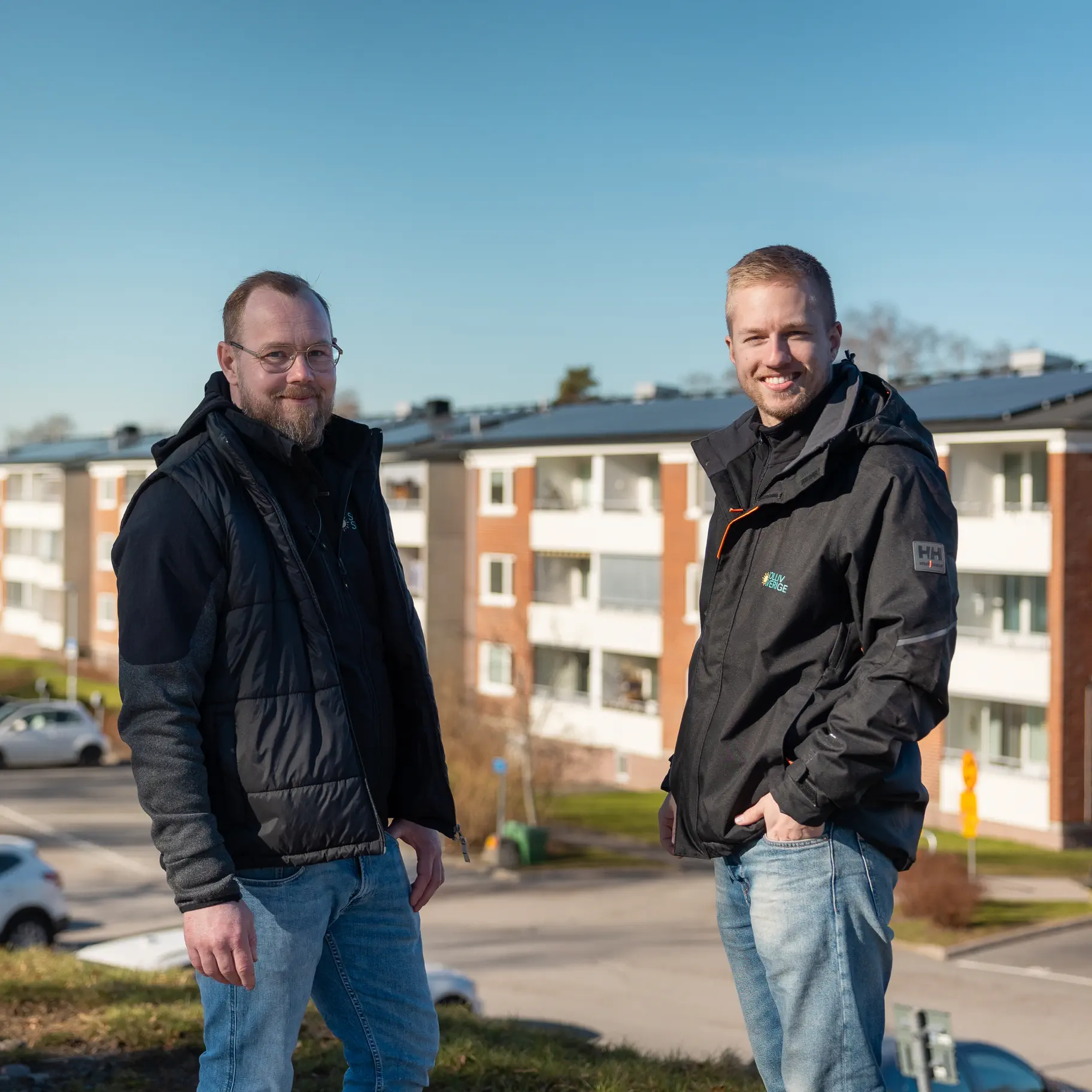 Johan Lundin och Simon Myrving från Solliv Sverige med ett flerbostadshus med solpaneler i bakgrunden.